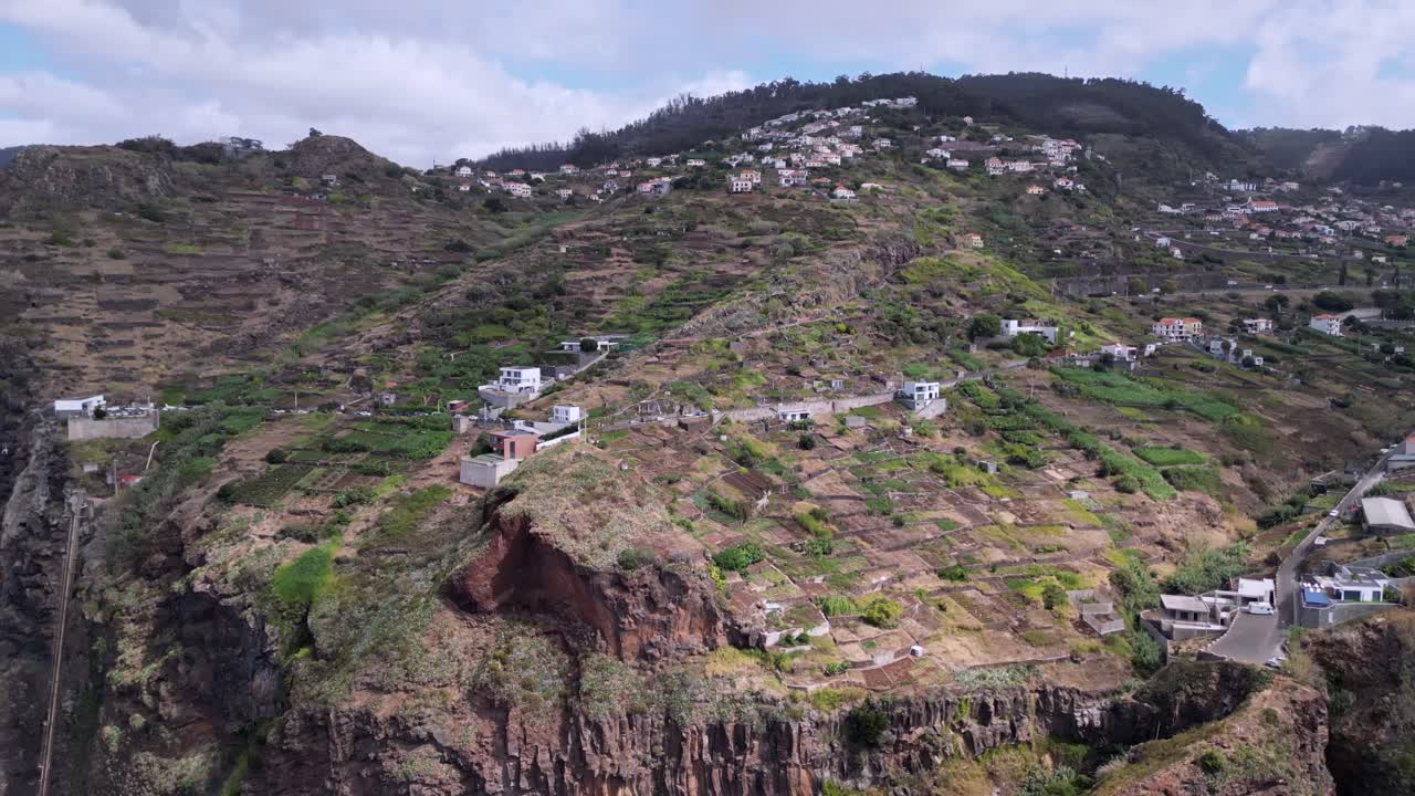 Aerial view of Madeira’s terraced hillsides, lush vegetation, dramatic cliffs, scattered houses, and winding roads under a cloudy sky.