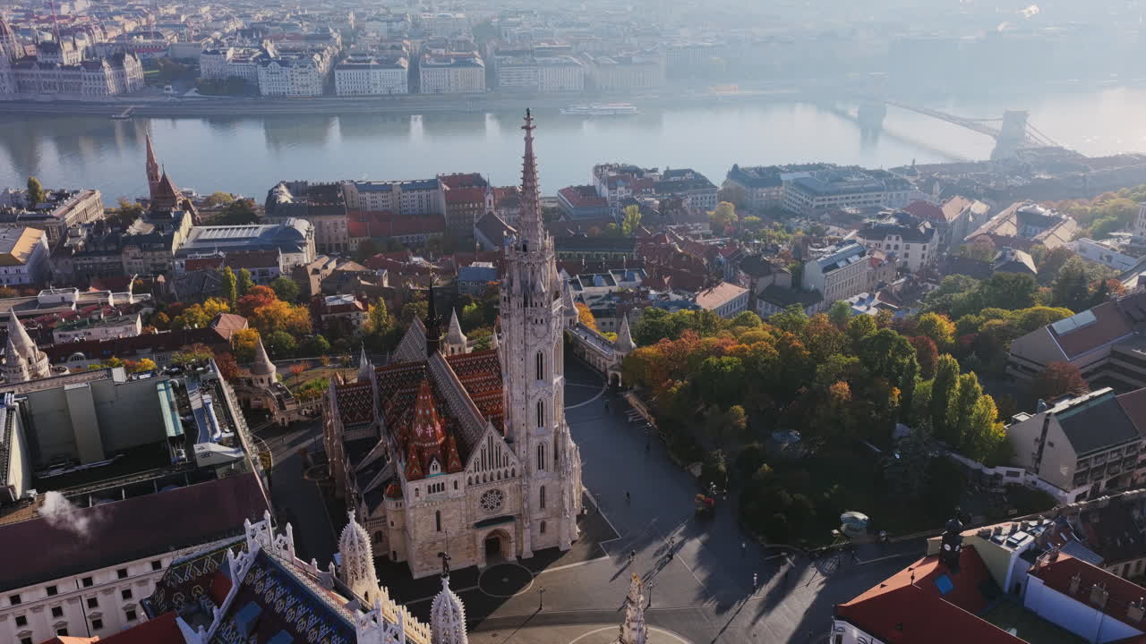La guglia alta e il tetto colorato con tegole della chiesa di Mattia si affacciano sul fiume Danubio, con il paesaggio urbano di Pest che risplende nella luce del mattino