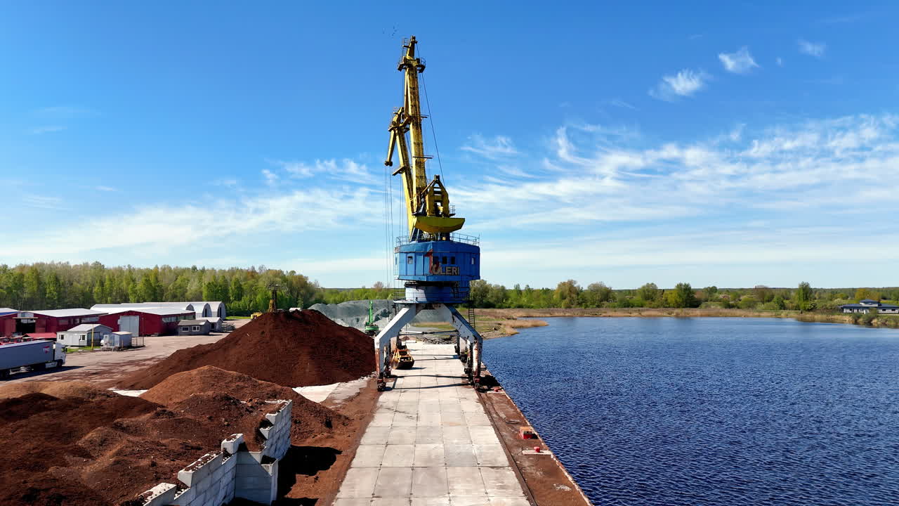 Tall yellow industrial crane positioned on a riverside dock beside large piles of bulk material, overlooking calm blue water and surrounded by green trees under bright daylight