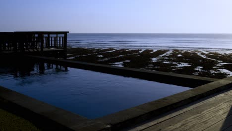 Toma-Estática-De-Una-Piscina-Al-Lado-De-La-Playa-Al-Final-De-La-Tarde-Con-Olas-Pacíficas-Rompiendo-Suavemente-En-La-Playa-De-Suluban-En-Bali-Durante-La-Marea-Baja-Con-El-Arrecife-Visible