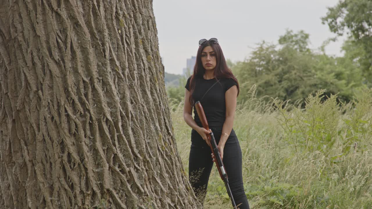 Donna nascosta dietro un albero che punta un fucile.