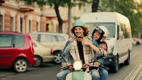 happy couple guy with long curly hair and in a denim jacket rides with his happy girlfriend in a plaid shirt who holds tightly to her boyfriend smiling broadly and looking at him while riding a moped on a city summer street