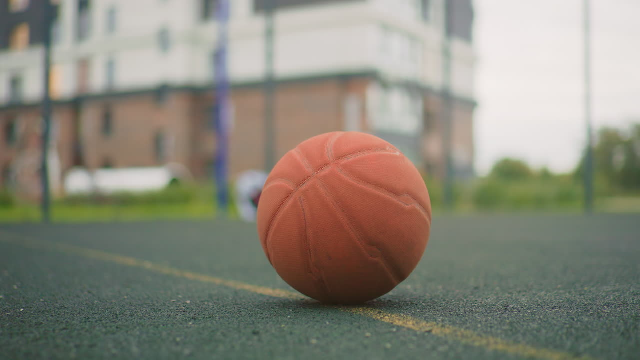 Bola basket oranye di lapangan basah dekat gedung apartemen, cahaya pagi dengan tetesan hujan di karet bertekstur, garis tengah berwarna kuning mengarah ke taman bermain yang buram dan mobil yang diparkir, close-up sudut rendah dengan kedalaman dangkal