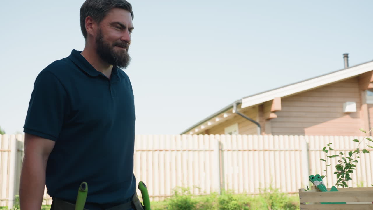 Young gardener tosses trowel, slides tool into apron belt on waist while gazing into distance near blue drum and wooden crate, house and fence behind, sunny backyard