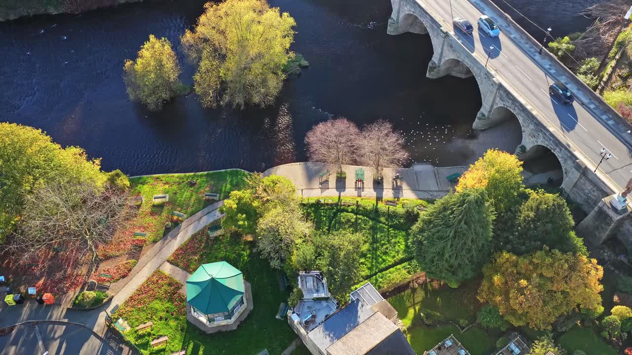 Aerial scene with Riverside Bandstand, mature urban park, historic arched bridge and tranquil river in Wetherby, Yorkshire. Autumn foliage, benches, and pathways visible in golden sunlight
