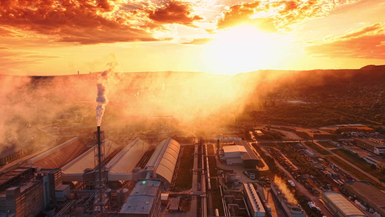 Hoge fabrieksschoorsteen die rook uitstoot in een heldere zonsondergang. Industriële rook stijgt op uit een metalen constructie tegen een feloranje zon en bewolkte lucht.