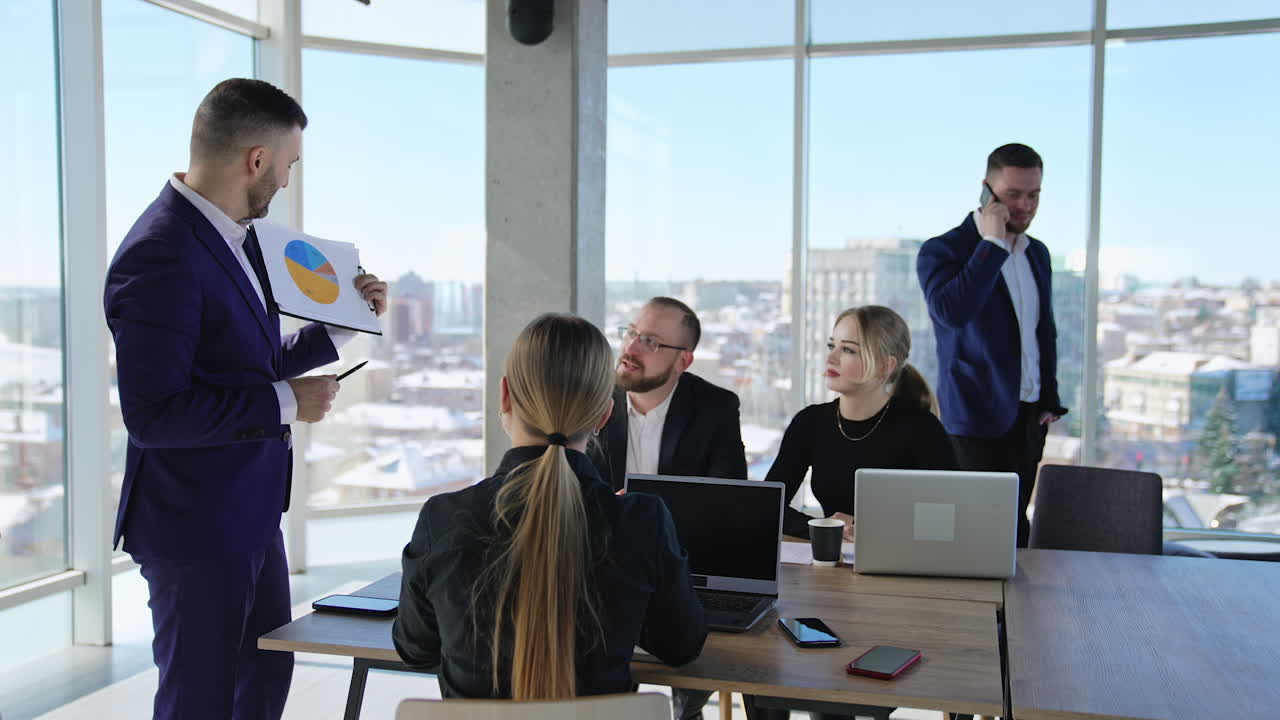 Office team having meeting sitting at desk and listening to their speaker. Man near the window speaking on the phone at backdrop.