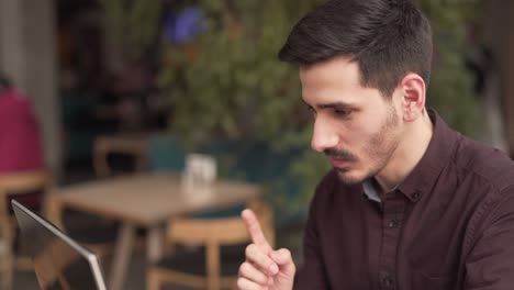 young man is sitting in a cafe in front of the laptop and talking with somebody in a video chat