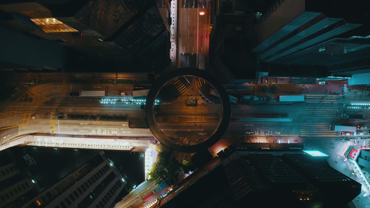 Drone shot traveling downward in the middle of a city toward a circle shaped crossroad during the night. the traffic is heavy with bus and cars