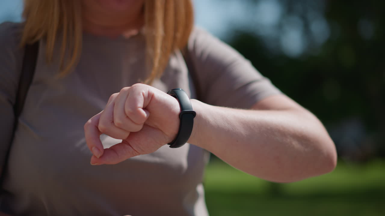 Close up of student checking time on smartwatch outdoors during bright sunny day, fingers adjusting screen under natural light, blurred green background