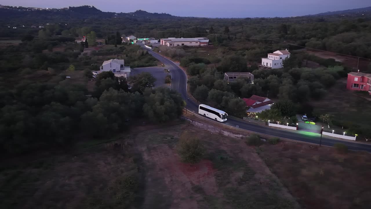 Aerial drone footage following a white tourist bus as it drives along a winding country road in Corfu, Greece, passing cars and houses at dusk