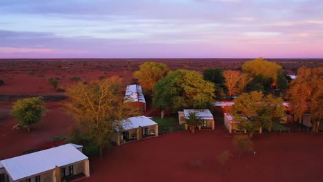 Drohne-Fliegt-Rückwärts-über-Eine-Lodge-Mitten-In-Der-Afrikanischen-Wüste-Mit-Vielen-Kleinen-Wohnungen-Und-Grünen-Anlagen-Im-Sonnenuntergang