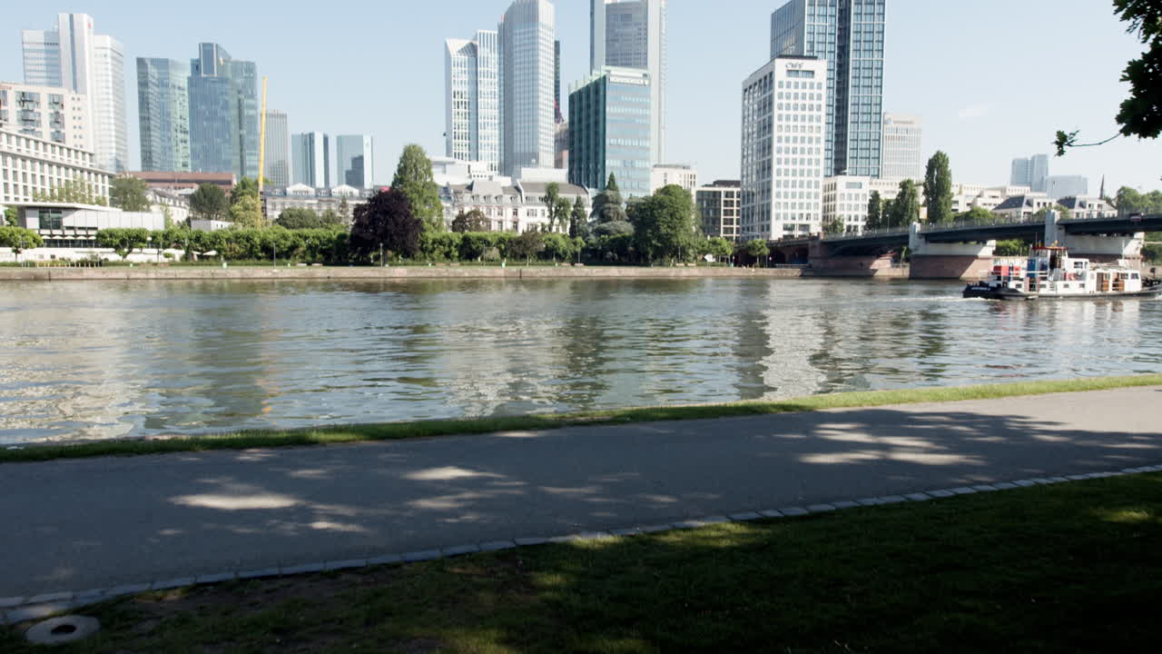 High-resolution video of Frankfurt's skyline and the Main River under a cloudless sky, shot in a wide angle from the shaded riverbank, showcasing serene city views