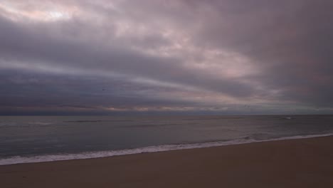 Sea-Waves-Splashing-On-Sandy-Shore-Under-Overcast-Sky-With-Birds-Flying