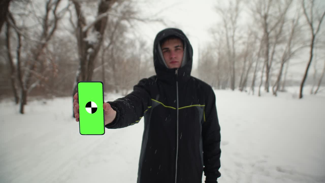 Caucasian man in black jacket with hood is standing on winter street and holding phone with green screen in his hands. Concept of winter pastime. Mockup