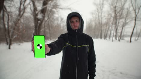 caucasian man in black jacket with hood is standing on winter street and holding phone with green screen in his hands. concept of winter pastime. mockup