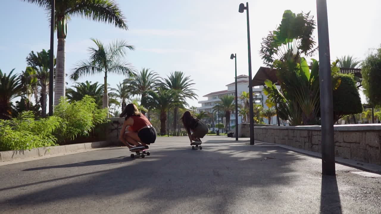 Embraced by palm trees and the sunset's hues, two young Hispanic women leisurely skateboard on island paths, their joyful activity portrayed in slow motion. A display of happiness and well-being