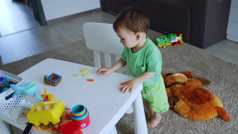 adorable toddler standing near the white desk. beautiful kid puts tiny toys into the colorful cups. top view.