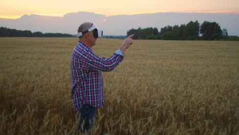 senior adult farmer in a virtual reality helmet in a field of grain crops. in the sunset light an elderly man in a tractor driver uses virtual reality glasses. vr technologies and modern agribusiness