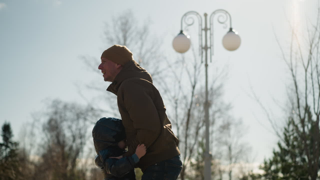 아빠는 행복하게 아들을 들어 올리고, 둘 다 청바지를 입고 있고, 아이는 한 재을 입고, 베니를 입은 남자가, 나무와 전기 기둥이 배경에 있습니다.