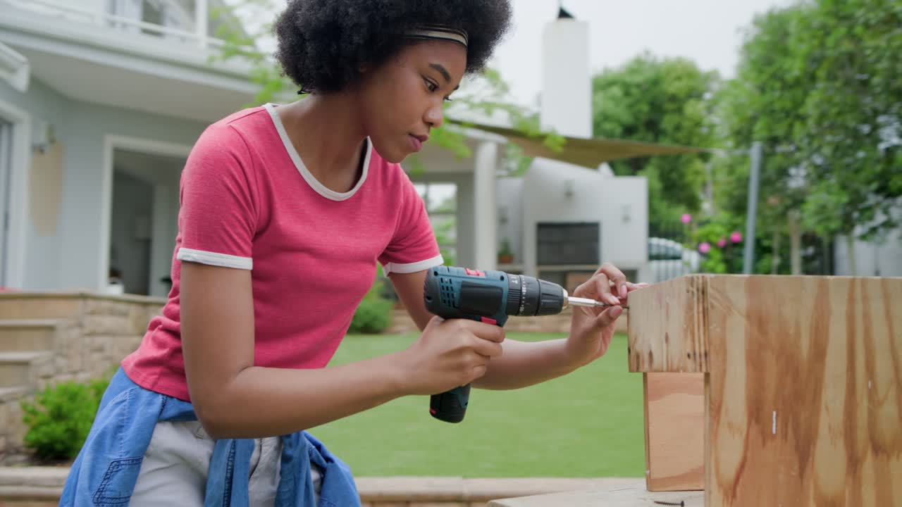 donna afroamericana che controlla il telefono, usando un trapano per fissare una scatola di compensato su una panca da patio in una maglietta rosa
