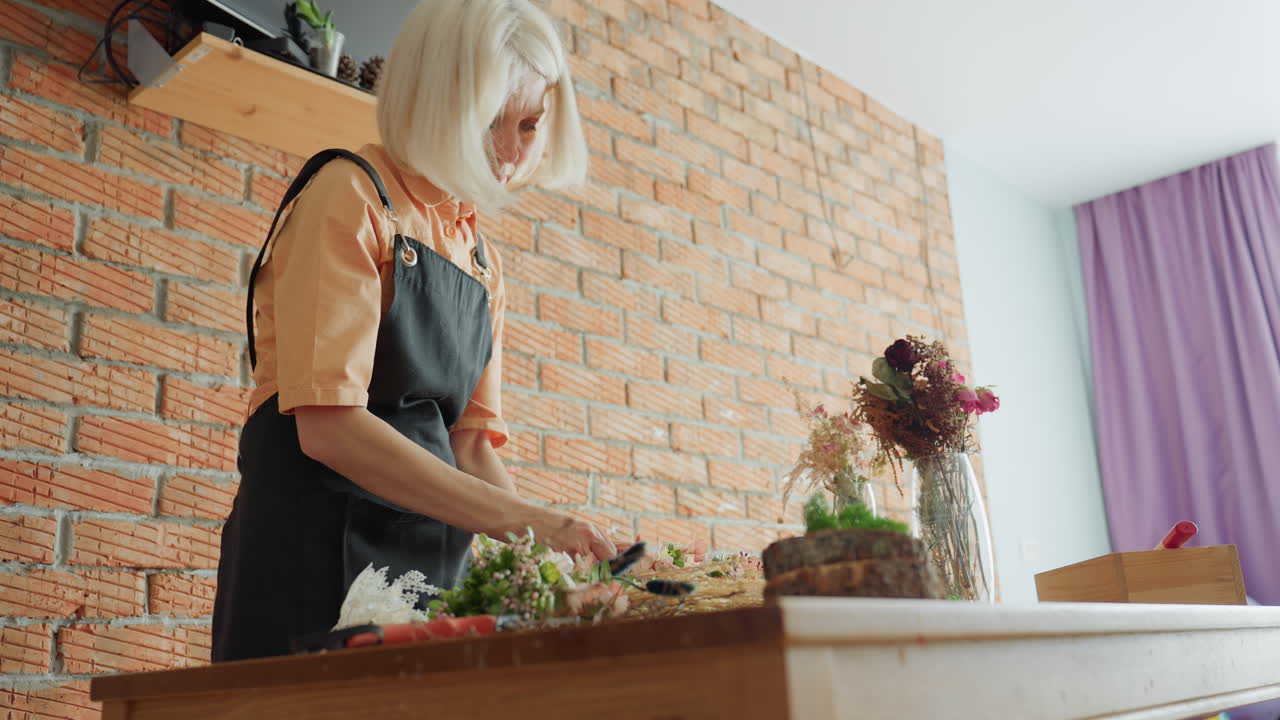 Eine blonde Frau mit schwarzer Schürze, die an einem Holztisch mit Werkzeugen, Pflanzen und Vasen auf einem rustikalen Kranz Blumen arrangiert und vor einer Backsteinmauer das florale Designhandwerk in einem gemütlichen, kreativen Arbeitsbereich im Innenbereich vorführt.