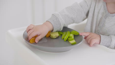 gros plan d'un bébé méconnaissable prenant un segment de clémentine de l'assiette dans une chaise haute