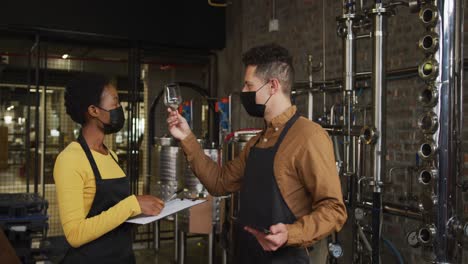 Diverse-male-and-female-colleague-in-face-masks-at-gin-distillery,-checking-product-and-making-notes