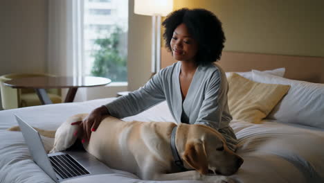 woman petting her labrador dog while working on a laptop in a cozy hotel room