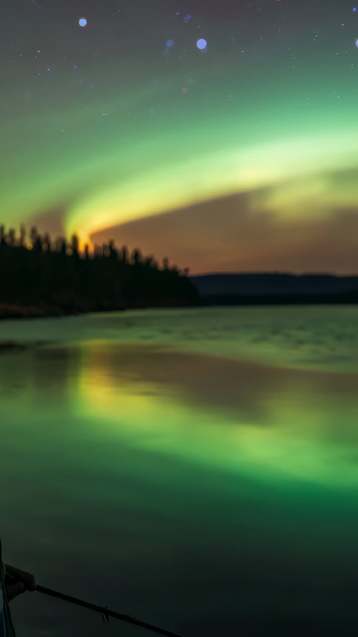 Vertical video: Entering, man holding rod, wearing beanie, fishing at lake under aurora, copy space