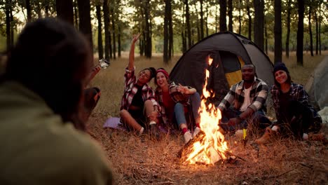 un hombre moreno con una chaqueta verde toma fotografías de excursionistas en grupo con camisas a cuadros cerca de un fuego. un hombre toca la guitarra y la gente posa en el marco cerca de una tienda en un bosque de otoño verde y amarillo