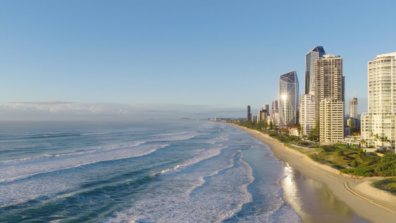 Aerial footage captures Gold Coast's beachfront skyline with high-rise buildings, ocean waves, and clear skies in bright daylight