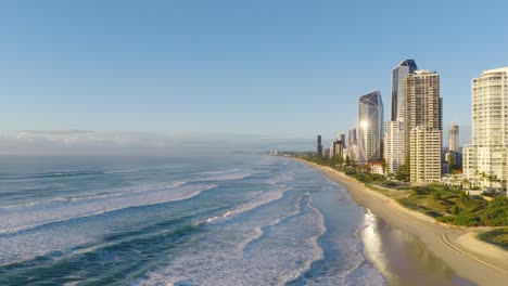 aerial footage captures gold coast's beachfront skyline with high-rise buildings, ocean waves, and clear skies in bright daylight