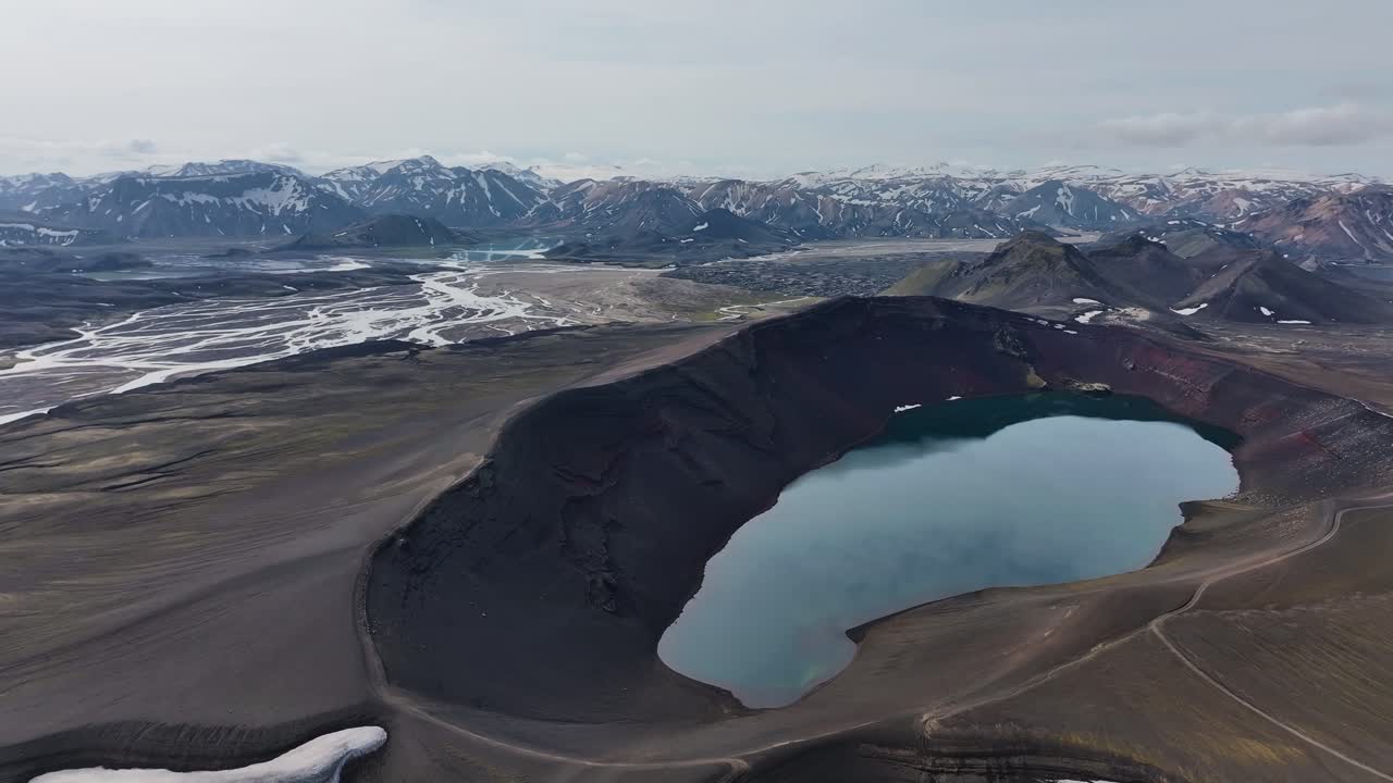 Slowing orbiting around a blue volcanic crater lake in the highlands of Iceland with a snow-capped mountain range in the distance