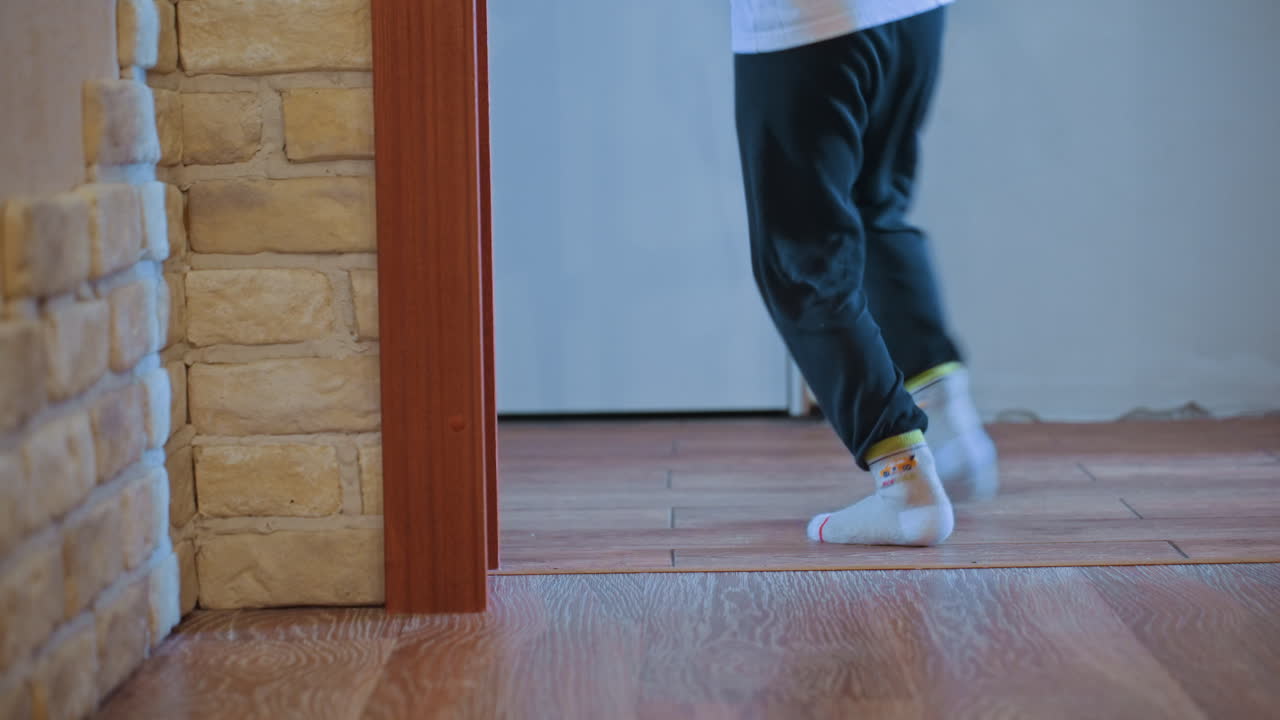 Son approaches kitten at doorway hardwood floor, sneakers and small hand entering frame, playful approach turns into gentle greeting, kitten watches from bricktrimmed jamb, ambient footsteps
