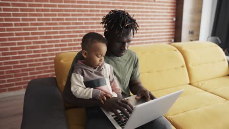 Joven-Padre-Afroamericano-Enseñando-A-Un-Niño-Pequeño-Enfocado-A-Usar-Una-Computadora-Portátil