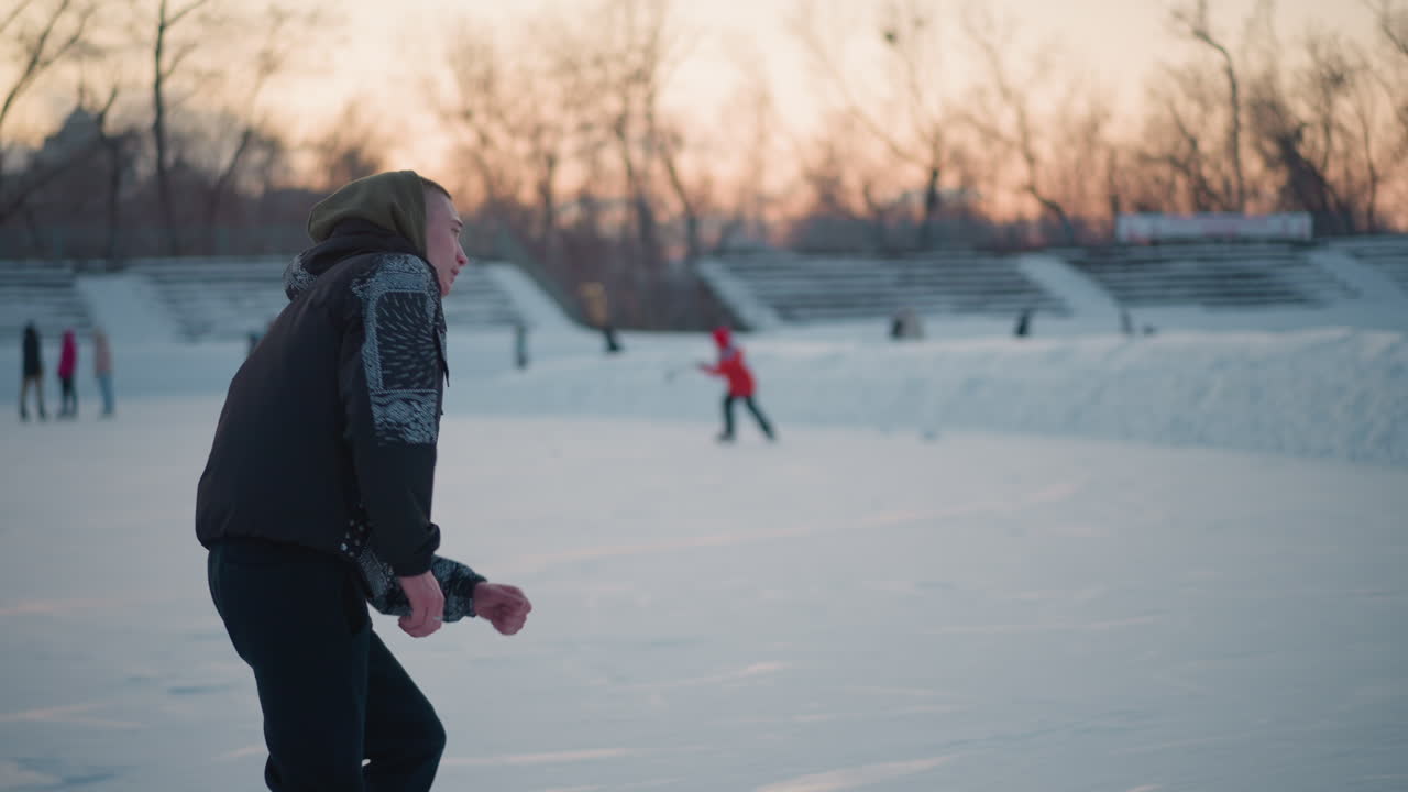 Young boy gliding on ice rink smoothly wearing winter jacket and hood with residential buildings and bare trees in background under sunset sky with distant skaters in snowy atmosphere
