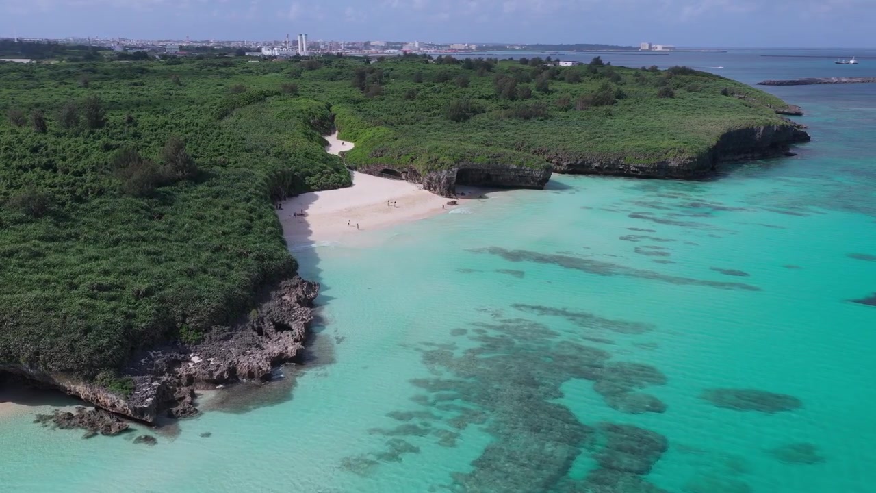 Inquadratura orizzontale con movimento di carrello in avanti verso sinistra, rivolta a destra, sopra la spiaggia sabbiosa, la costa lussureggiante e le acque turchesi.