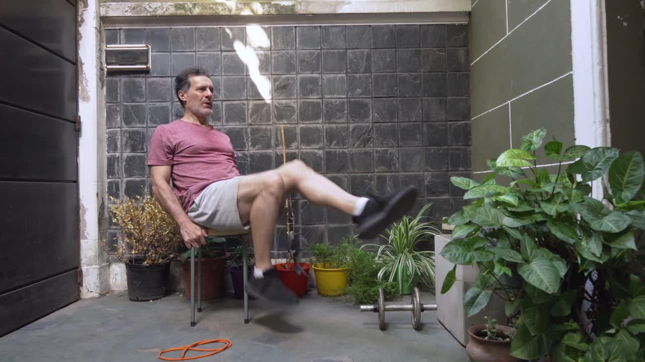 Mature man with glasses uses a chair in a domestic outdoor setting to perform a seated bicycle exercise, targeting his abdominal and oblique muscles