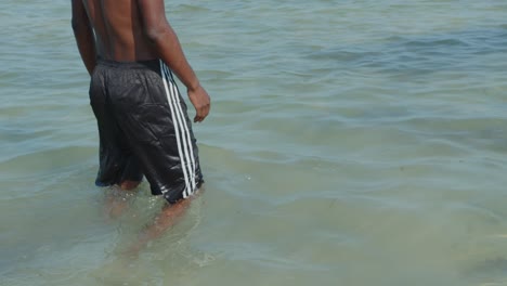 un hombre negro vestido con pantalones cortos negros camina hacia el océano con el agua hasta la cintura
