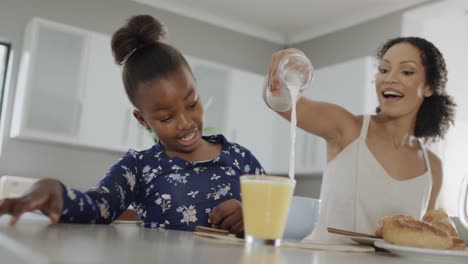 happy african american mother and daughter eating breakfast in kitchen, slow motion