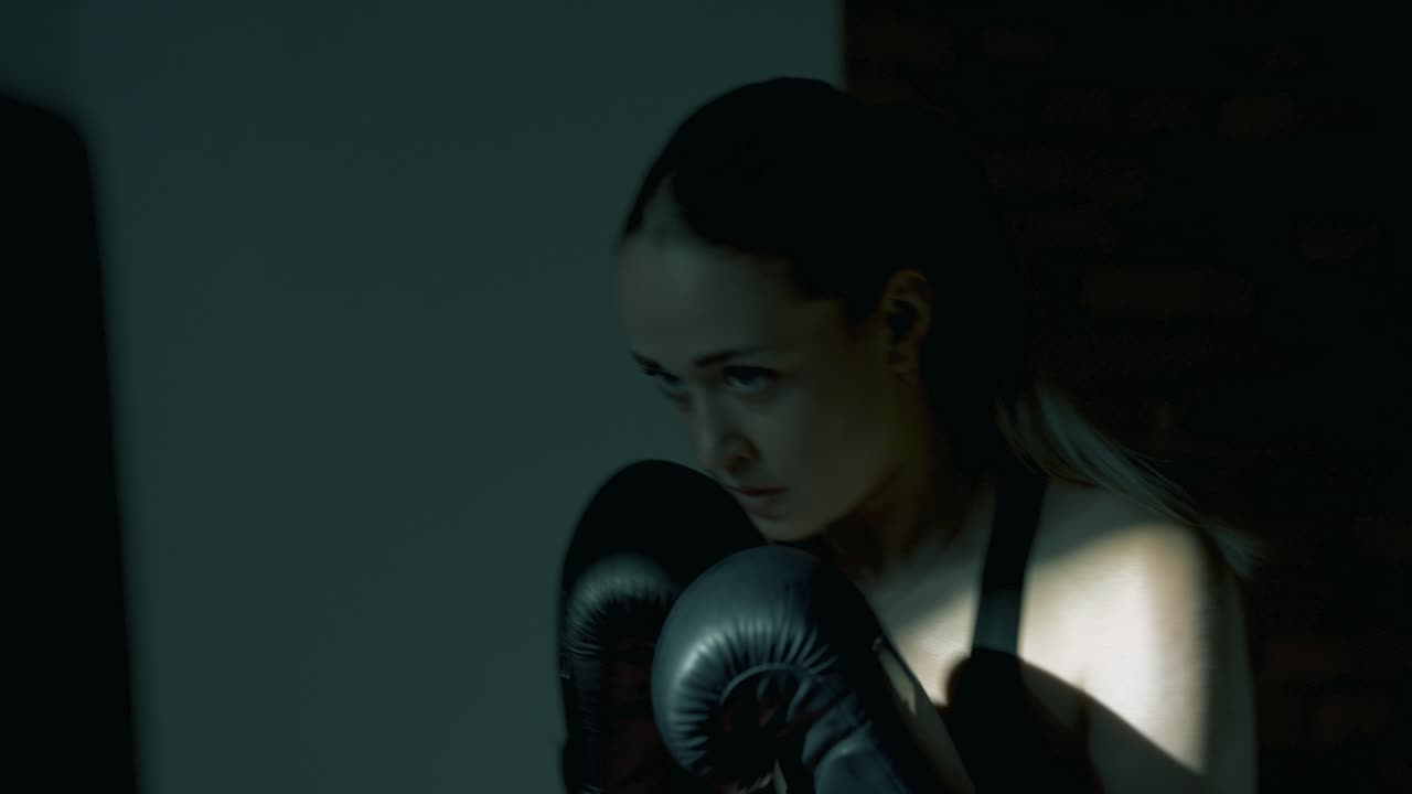 Profile shot of blonde woman boxing a heavy bag with large black gloves. Camera briefly moves away, showing punches through the bag, then returns to profile. Dramatic gym light and motion