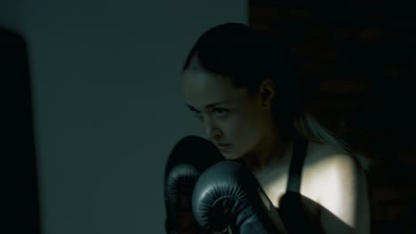 Profile shot of blonde woman boxing a heavy bag with large black gloves. Camera briefly moves away, showing punches through the bag, then returns to profile. Dramatic gym light and motion