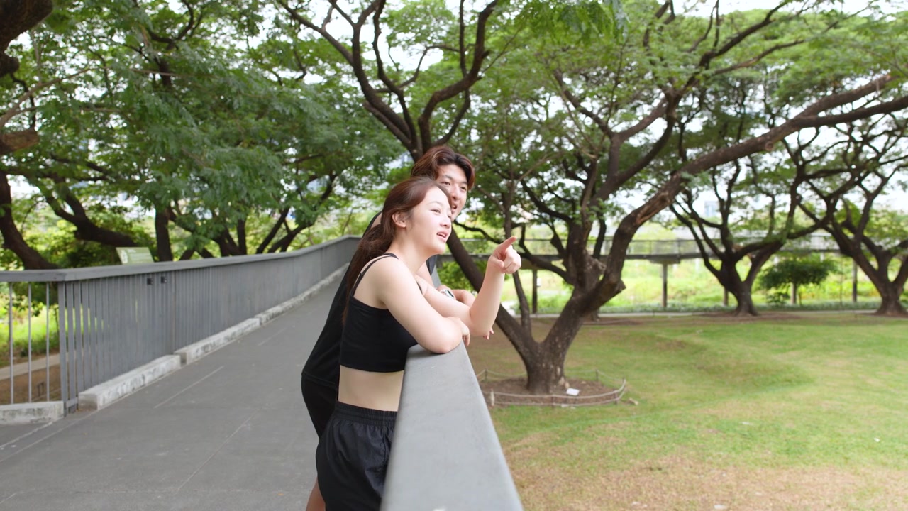 黒いスポーツウェアを着た若いタイ人カップルが、公園の手すりに寄りかかり、話したり指差したりしている。