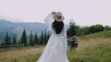 Rear view of a bride in the mountains. Girl in wedding dress and pink hat holding bouquet of flowers and looking on the beautiful mountain landscape.