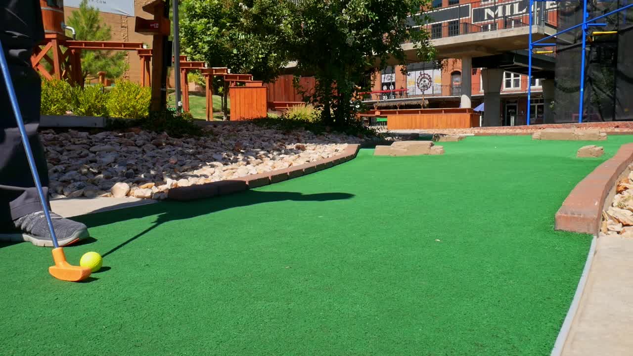 Playing mini golf with a yellow ball and orange putter on green astroturn on a sunny day - shot from the side