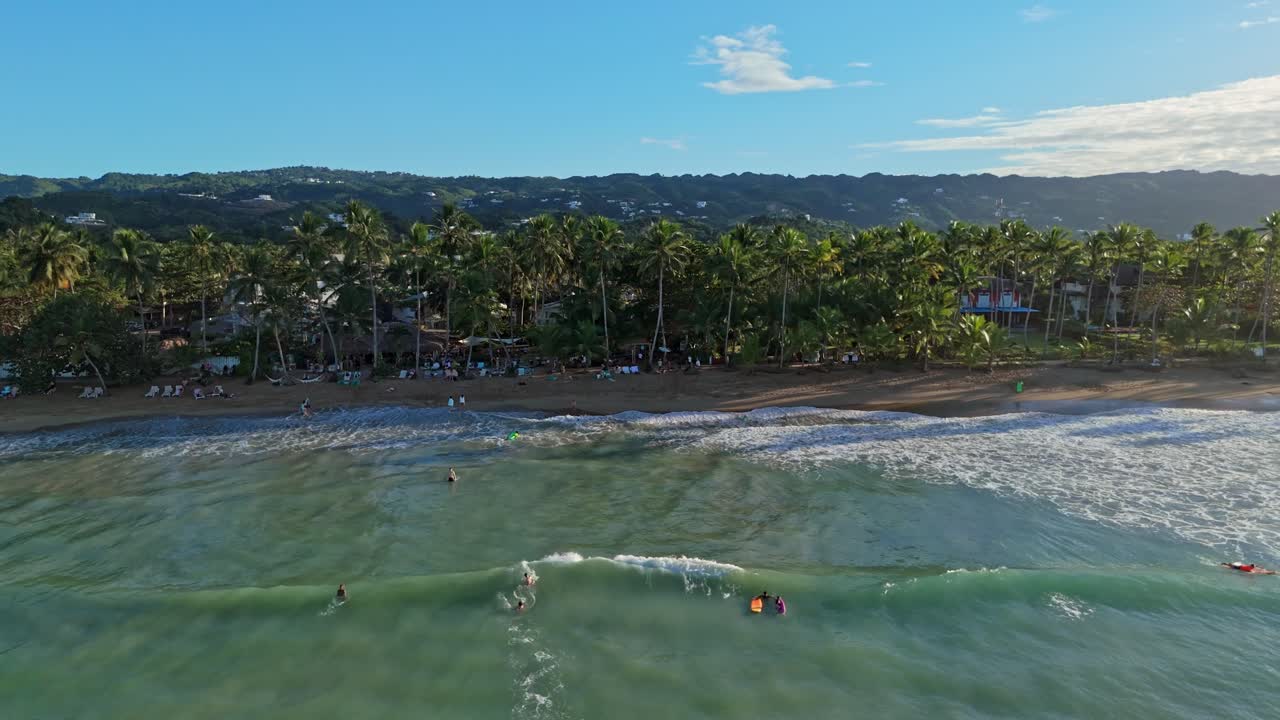 Swimming people in clear Caribbean sea during sunset. Palm trees at sandy Playa Bonita Beach during sunny day. Aerial lateral wide shot.