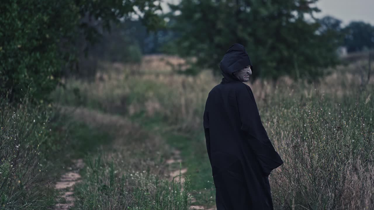 Backside portrait of death in nature. Dark figure in black costume out stretches its arms and goes away in the field in the evening. Scary nightmare ghost outdoors.