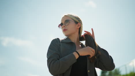 lady wearing black crop top under grey shirt gently holds her hair while looking away, she stands outdoors on a sunny day, her hair fluttering slightly in the wind