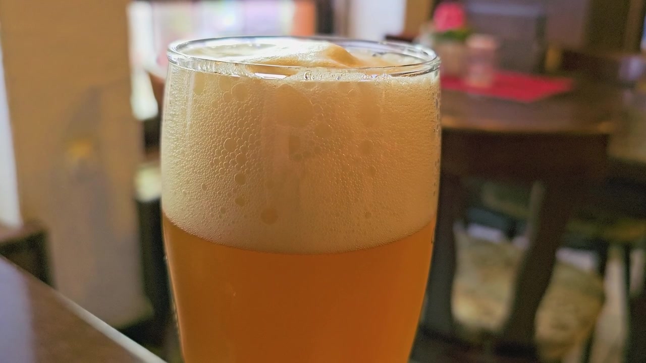 Nahaufnahme eines kalten Bierglases mit dickem Schaum, goldener Farbe, Bläschen, Kondensation und verschwommenem Restaurant-Hintergrund, ideal für Aufnahmen in Kneipen, Brauereien, Getränke und zwanglosen Restaurants.